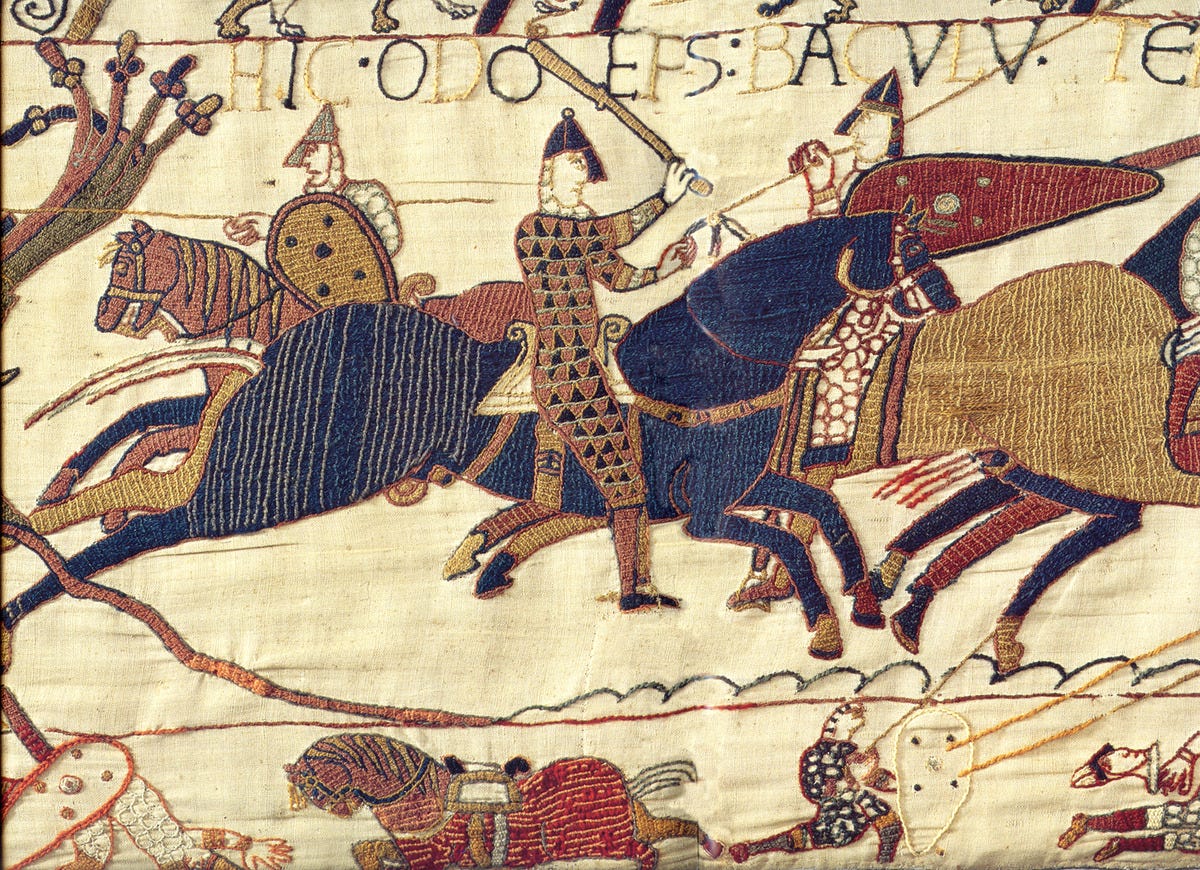 A detailed scene from the Bayeux Tapestry, a medieval embroidered cloth depicting the Norman Conquest of England in 1066. This section shows armored knights on horseback in battle, with one central figure wielding a club and labeled in Latin as “HIC ODO EPS BACULU(T) TENENS” (Here Bishop Odo holds a club). The horses are shown in motion, and soldiers engage in combat below. The stitching is bold and colorful, with geometric patterns on clothing and armor, emphasizing action and hierarchy. A scene from the Bayeux Tapestry depicting Bishop Odo rallying Duke William's army during the Battle of Hastings in 1066, via [Wikimedia](https://en.wikipedia.org/wiki/Bayeux_Tapestry#/media/File:Odo_bayeux_tapestry.png)