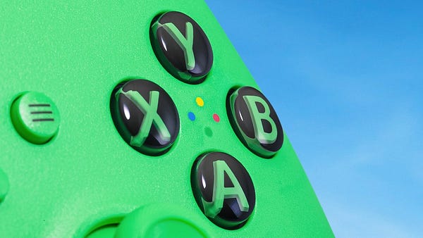 An extreme close-up of the Velocity Green Xbox controller shows the ABXY buttons and Start button against the main green of the controller. The ABXY buttons themselves are black, while the ABXY letters inside match the same green. The start button matches the same green color as the controller, with a black hamburger menu icon in the center. In the center of the ABXY buttons, small green, red, blue, and yellow dots indicate which letter button is which. The controller floats against a bright, beautiful blue sky background.