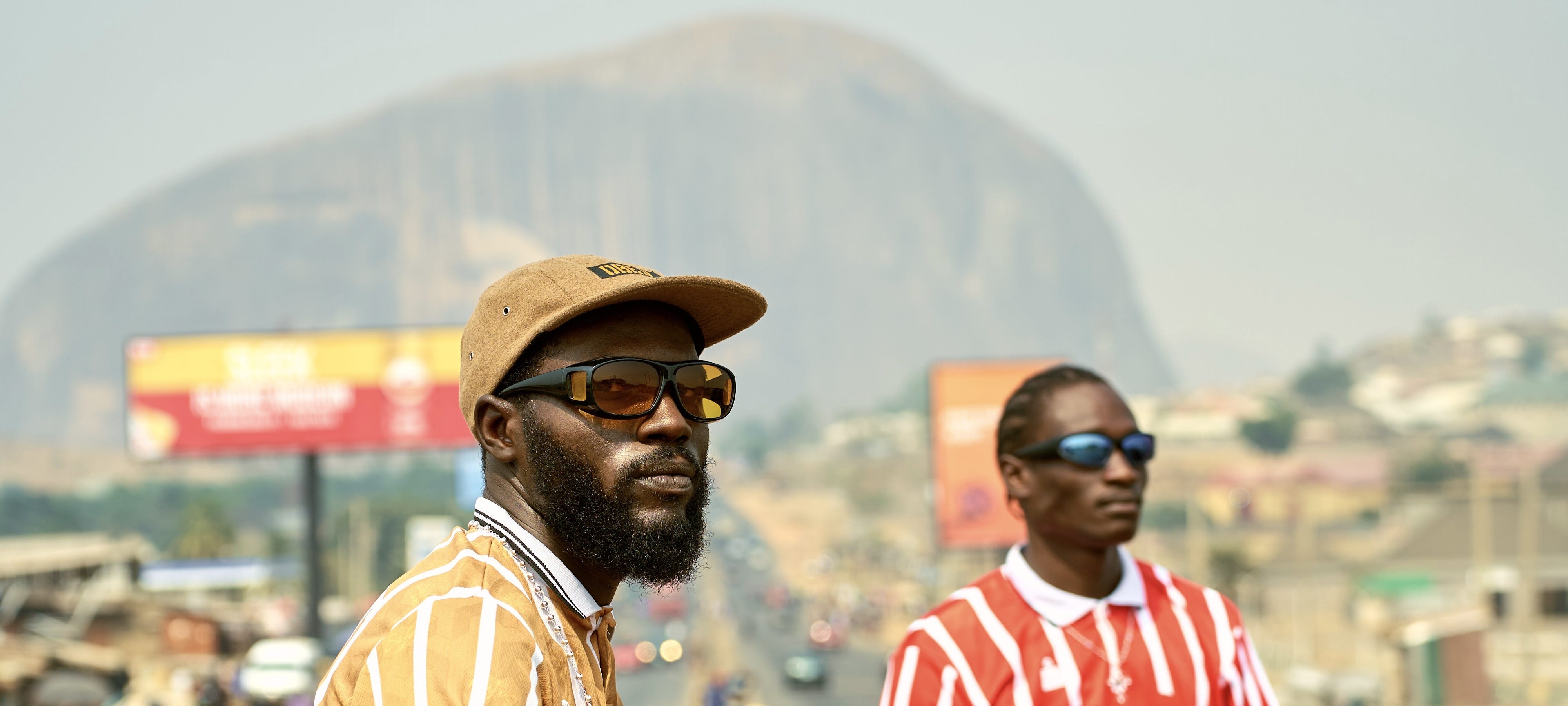 Two young men in striped football jerseys and sunglasses on a highway median with Zuma Rock looming behind