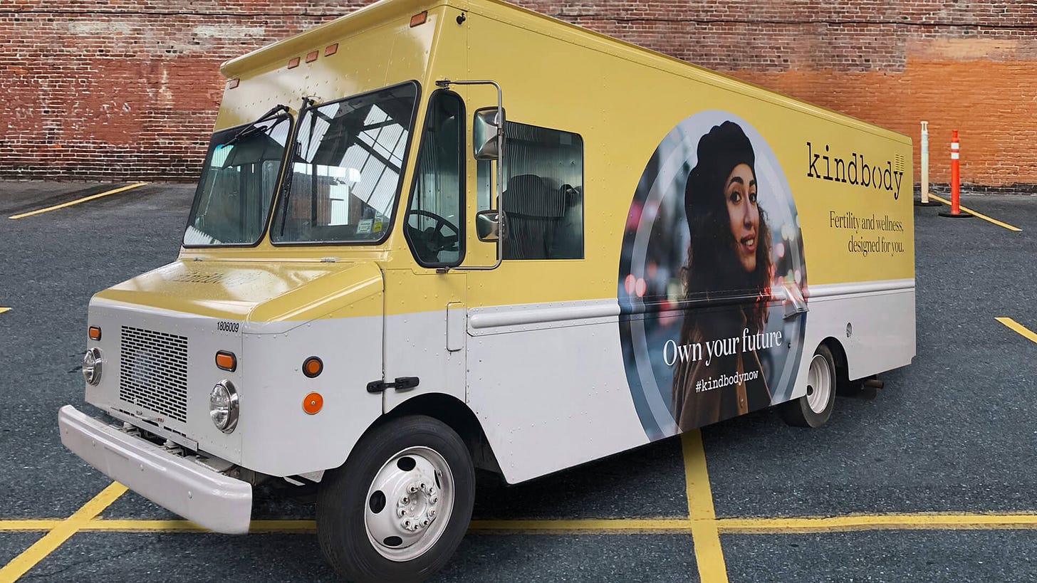 Minibus giallo e bianco della clinica Kindbody, di cui si legge il motto sotto il logo: “Fertility and wellness, designed for you”. Sulla fiancata del bus, la foto di una ragazza sui 30 anni con lunghi ricchi neri che guarda l’obiettivo sorridente. Sotto di lei la scritta “Own your future #kindbodynow”. 