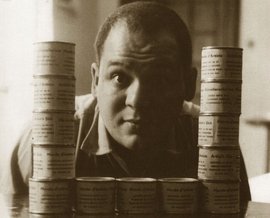 b/w shot of Piero Manzoni with 13 tins of Merda d’Artista stacked and laid out to form a squarish U-shape. He is peering through the gap in the middle