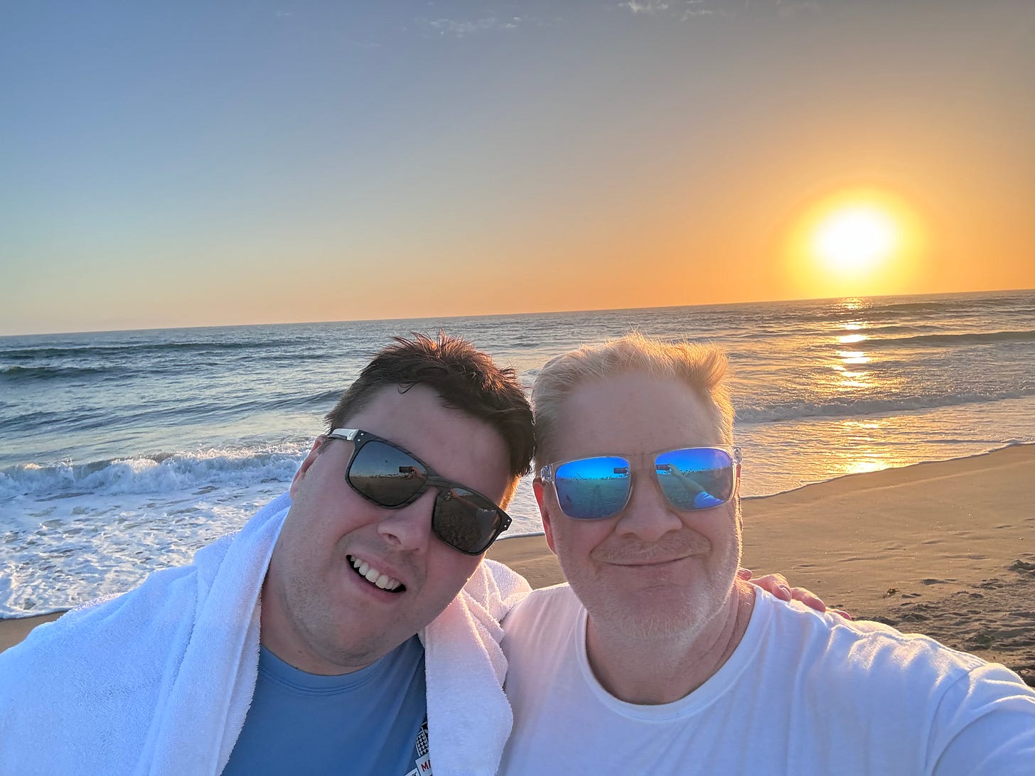 J.B. Handley and his son Jamie smiling in sunglasses on a beach at sunset