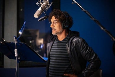 Photos of Naveen Andrews in a recording booth with a microphone above his head. To the right is a headshot of Wrenn Schmidt wearing a multi-colored striped shirt.