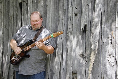 Eric Beaty playing guitar posing with Breedlove and Ibanez guitars wearing a NaNoWriMo winner shirt and posing with Smart Focus book series