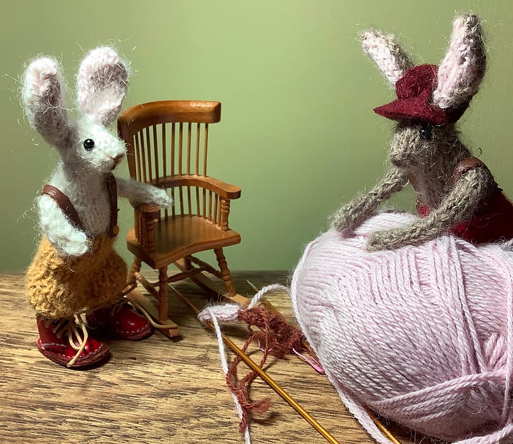 Delores and Bernard two knitted rabbits are with a ball of yarn looking at knitting needles with some stitches on.
