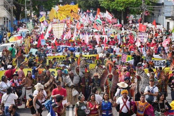 A colorful throng of people, some holding large flags and others wearing Indigenous clothing, gather in a street.