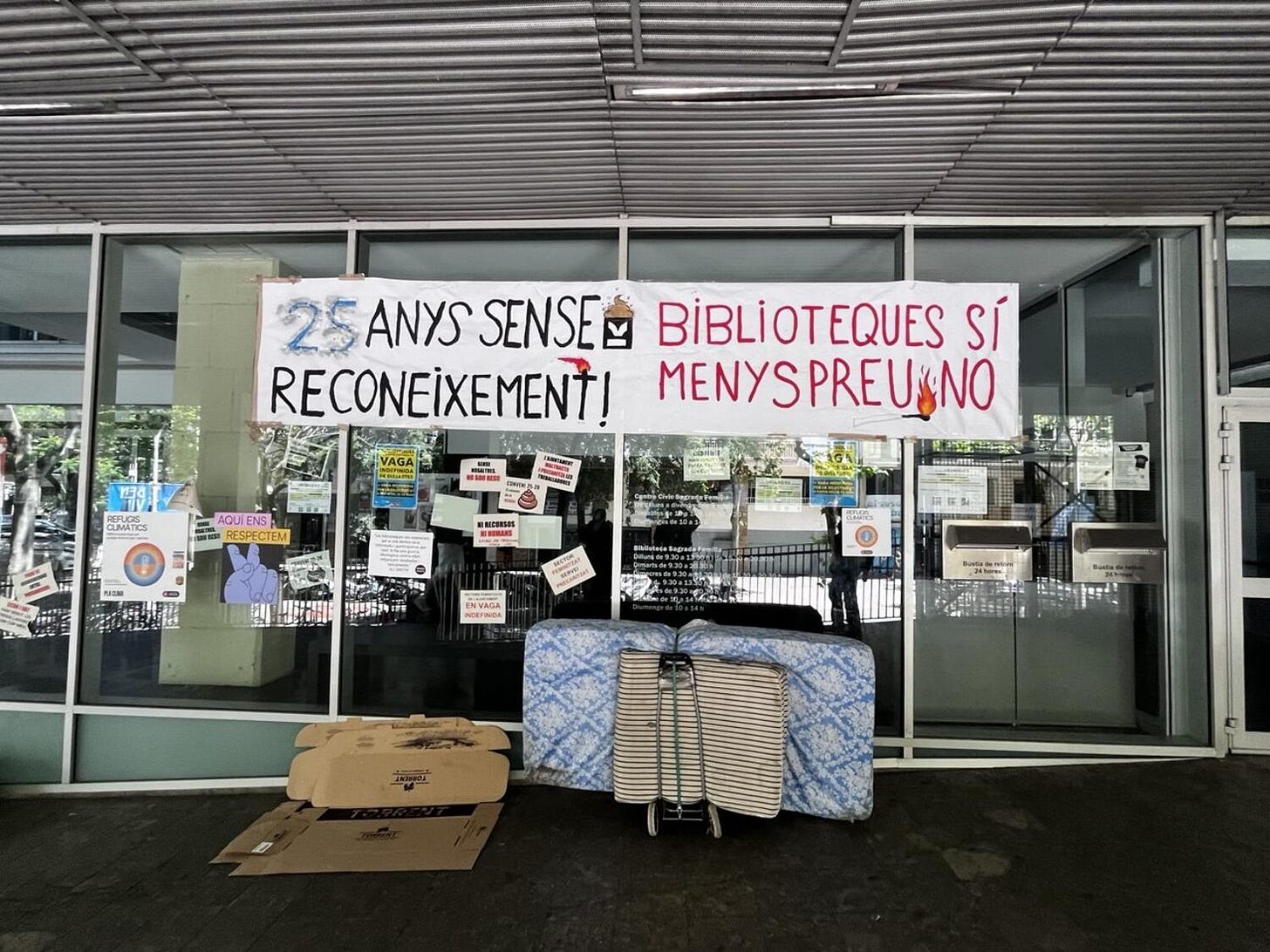 La facciata in vetro di una biblioteca di Barcellona ricoperta di adesivi: alcuni dicono che quello è un rifugio climatico della città, molti altri sono rivendicativi dello sciopero. In alto lo striscione in catalano "25 anys sense reconeixement! Biblioteques sì, menyspreu no". Davanti alle porte, un materasso e i cartoni che fungono da giaciglio per una persona senza tetto, non presente nella foto.