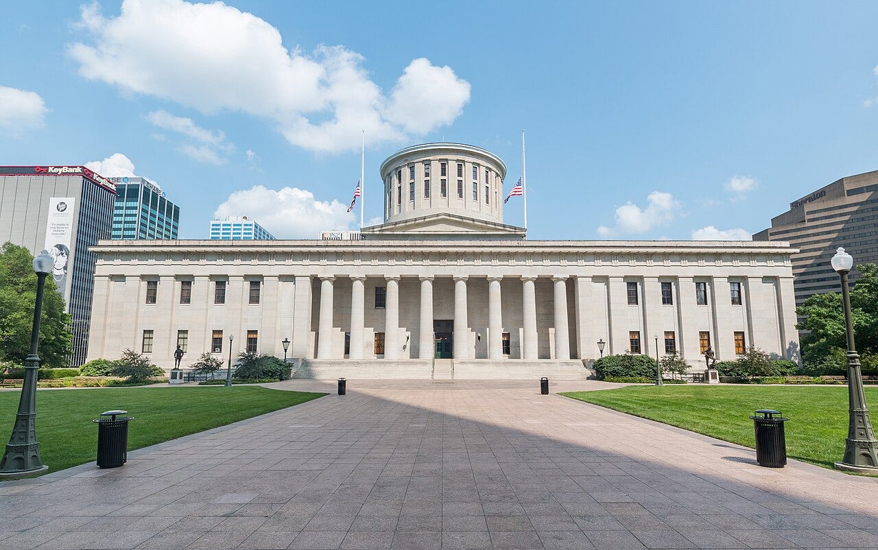 Ohio Statehouse exterior, Columbus — a neoclassical building with a colonnade of tall stone columns, a low rotunda dome, and three flags flying at half-staff, flanked by manicured lawns and city buildings in the background.