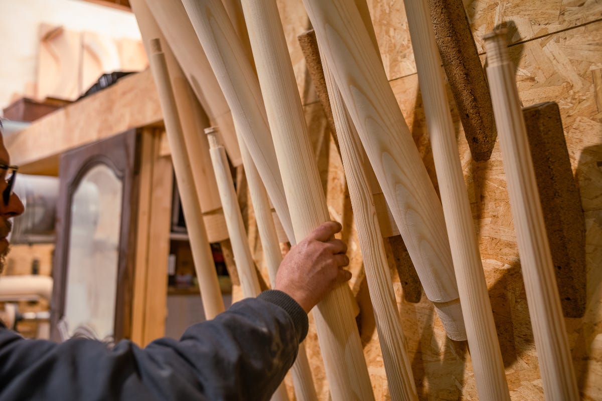 A person’s hand, selects a polished wooden tube from a rack of partially assembled alphorns.