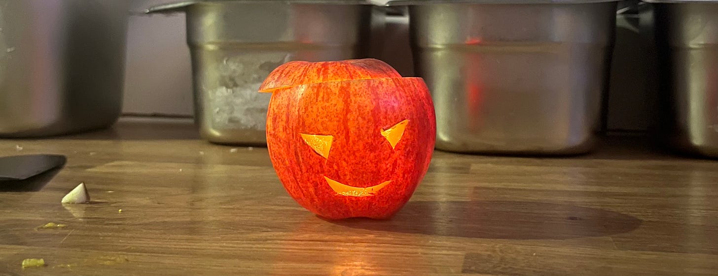 a photograph of an apple carved like a traditional Halloween lantern, against a background of gastros on a quite messy kitchen bench