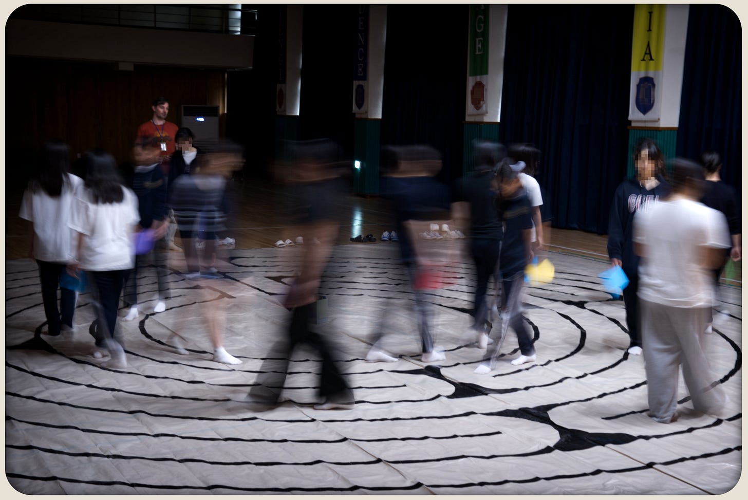 Students walk a taped labyrinth on a tarp in a dim auditorium; motion blur shows continuous movement, with some holding coloured papers.