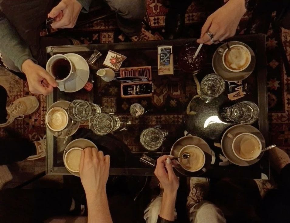 This may contain: four people sitting at a table with cups of coffee in front of them and one person holding a spoon This may contain: four people sitting at a table with cups of coffee in front of them and one person holding a spoon