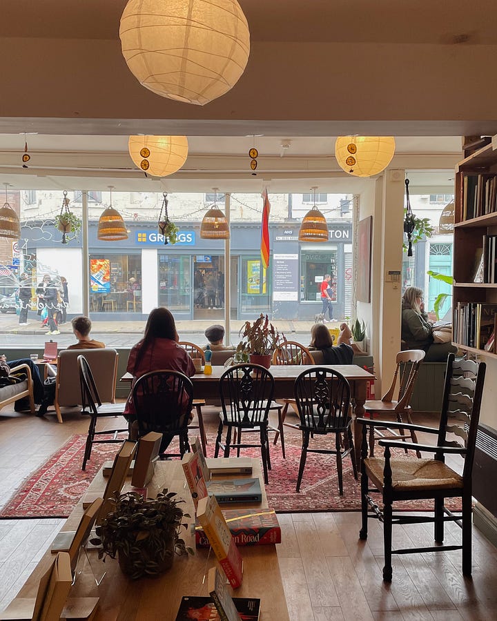 Image 1: Holding an almond croissant with a bite taken out, sitting cross-legged. Sand is visible below. / Image 2: The tide rolling in, gentle waves meeting the shore. / Image 3: Inside Tills & Company bookshop—books, tables, and people enjoying a slow moment. / Image 4: A coffee table in Tills with my book Ways of Seeing and a coffee cup resting on it.