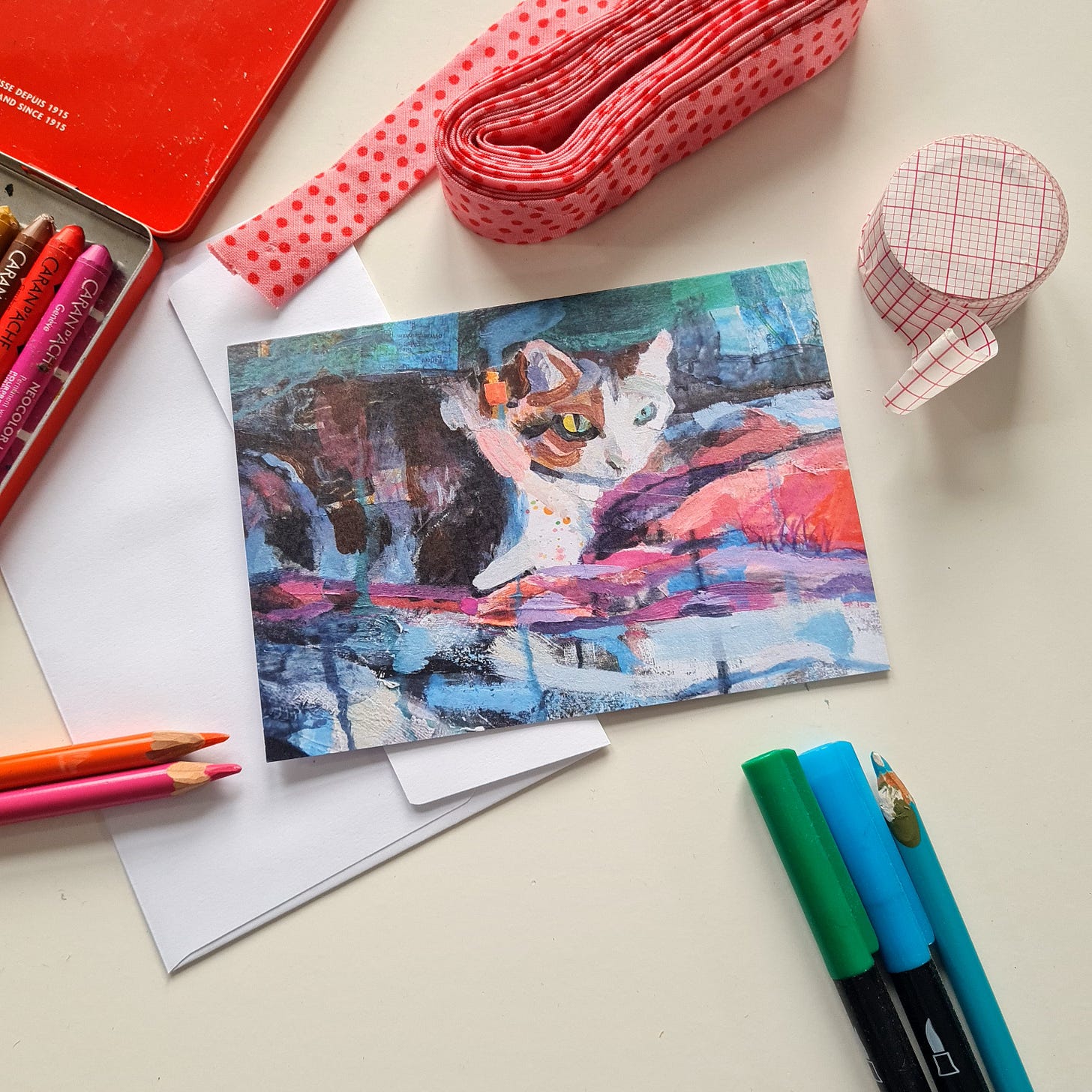 A greeting card with a painting of a cat on the front is on a white table surrounded by aty materials and stationery items. A greeting card with a painting of a cat on the front is on a white table surrounded by aty materials and stationery items.
