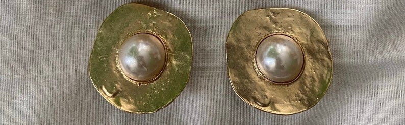 May include: Pair of gold-tone statement earrings. Each earring features a textured, irregular, round shape with a central, off-white pearl. The gold metal has a hammered or textured finish, giving it a slightly rustic look.  The pearls are smooth and glossy, providing a contrast to the matte gold. These earrings are a classic, elegant design with a modern twist.