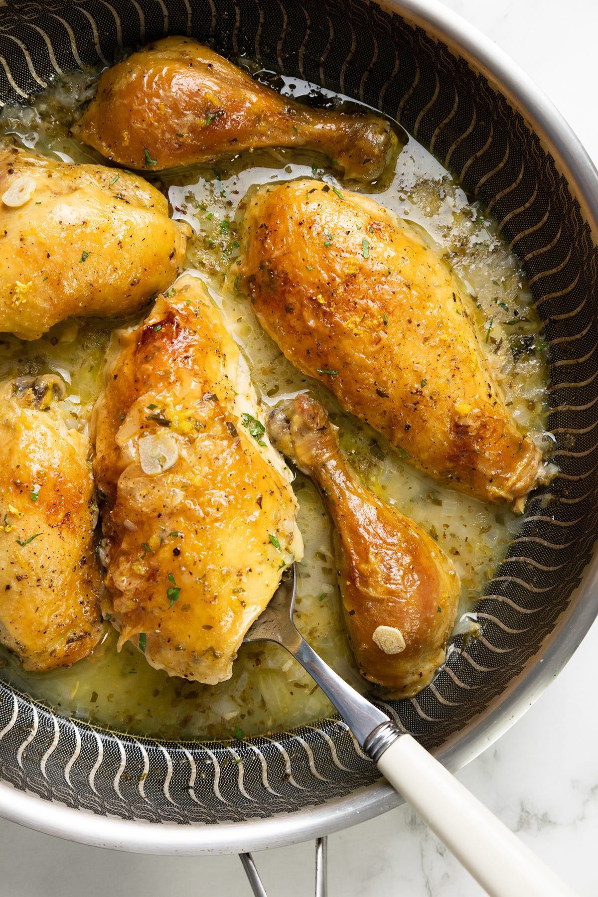 Golden-brown chicken pieces in a skillet with a lemony, herb-infused sauce, garnished with sliced garlic and fresh herbs. A serving spoon is lifting one piece.