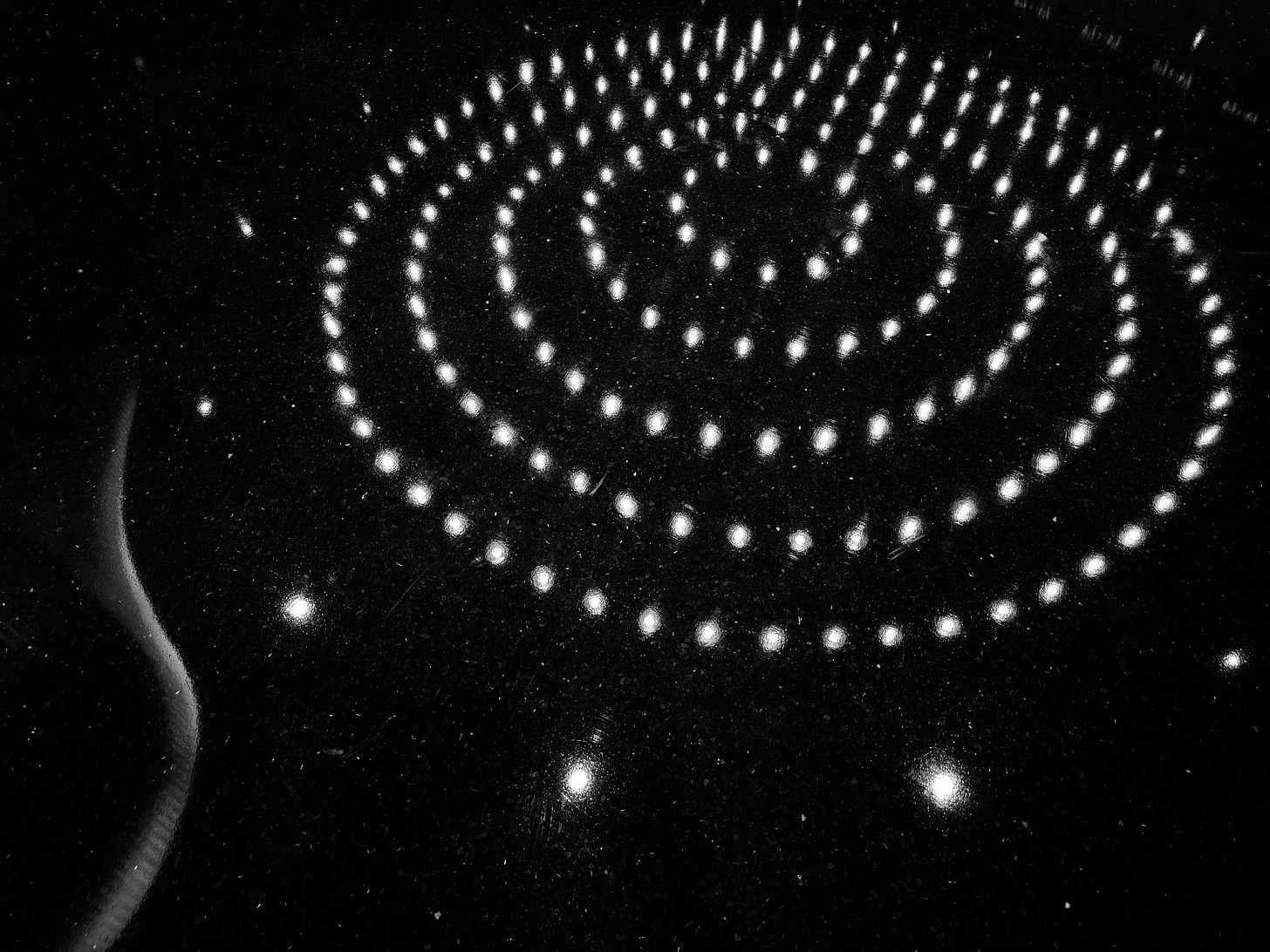 Per ChatGPT: The image is a black and white photograph showing a pattern of lights arranged in concentric circles. The lights appear to be small and bright against a dark background, giving the impression of being stars or possibly lights from a man-made structure reflected on a surface. The exact nature of the surface is unclear, but it could be water, glass, or another reflective material given the clear reflections and the way the lights diminish in intensity with distance. There is a swirled pattern that disrupts the concentric circles on the left side, adding an element of motion or distortion to the image. Per ChatGPT: The image is a black and white photograph showing a pattern of lights arranged in concentric circles. The lights appear to be small and bright against a dark background, giving the impression of being stars or possibly lights from a man-made structure reflected on a surface. The exact nature of the surface is unclear, but it could be water, glass, or another reflective material given the clear reflections and the way the lights diminish in intensity with distance. There is a swirled pattern that disrupts the concentric circles on the left side, adding an element of motion or distortion to the image.
