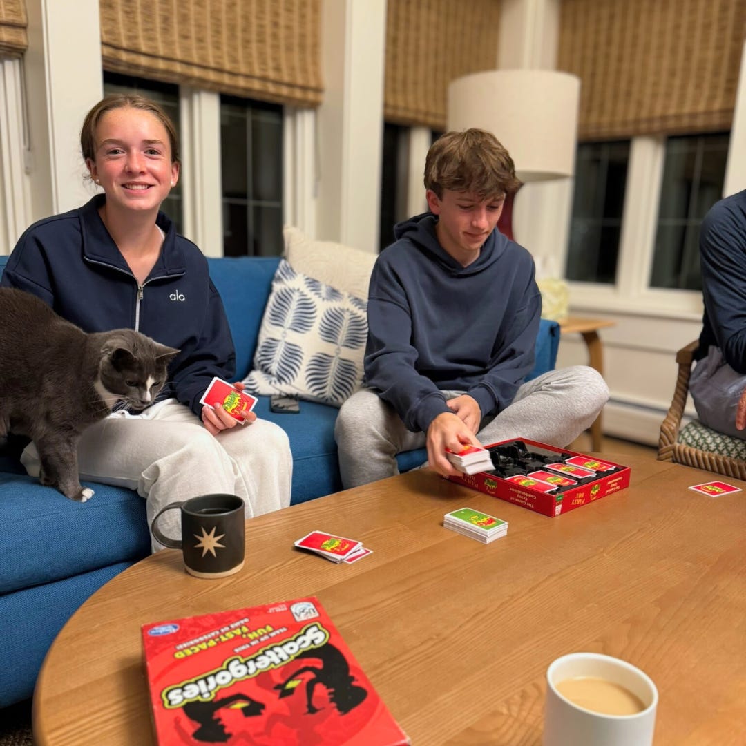 Marilee and William sit on a blue couch playing the card game Apples to Apples. A gray cat stands next to Marilee on the left, who is smiling at the camera and holding cards. William beside her is shuffling cards from the box on the table.