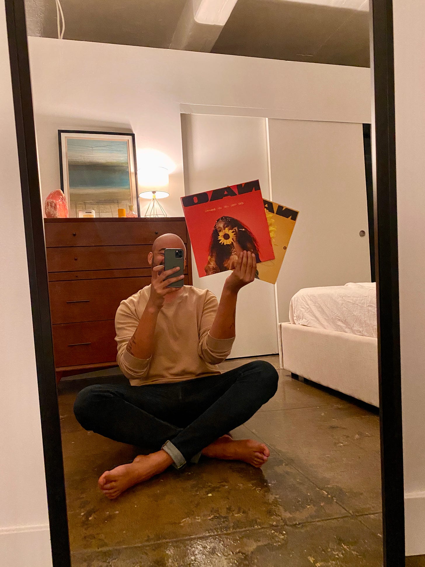 A mirror selfie of a man sitting cross-legged on a concrete floor, holding up two vinyl records by Nao. The setting is a minimalist apartment with a wooden dresser, a salt lamp, and a framed painting visible in the background.