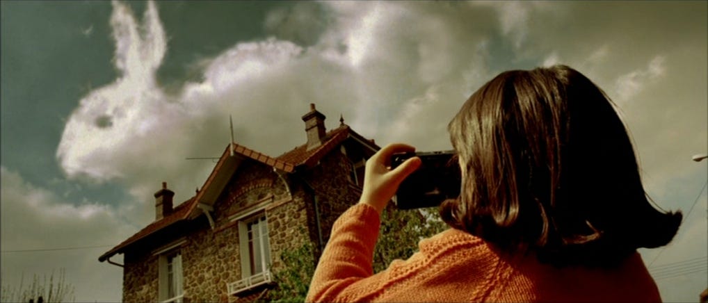 A film still from the movie Amélie in which we see the title character as a child. We see the back of Amélie's head and orange sweater as she takes a photograph of the sky behind her house. In the sky is a cloud in the shape of a rabbit.
