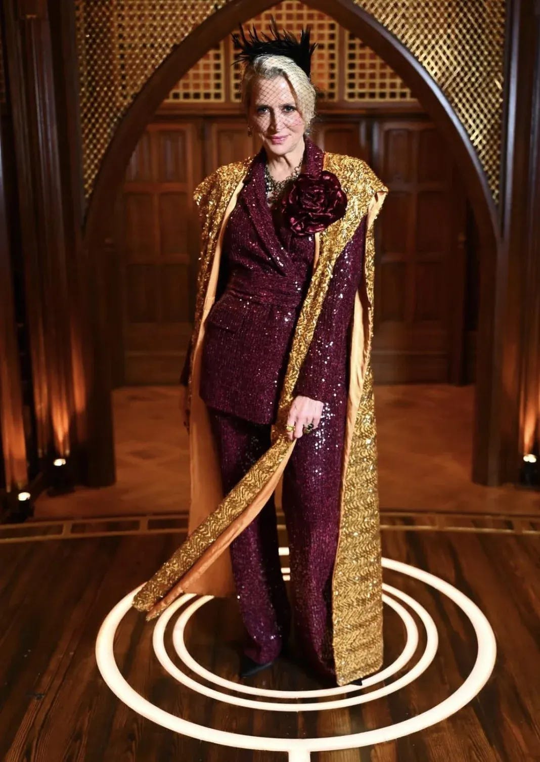 Dorinda in a burgundy sequined pantsuit with a floor-length gold embroidered cape, burgundy floral brooch, and a black veiled fascinator with feathers.