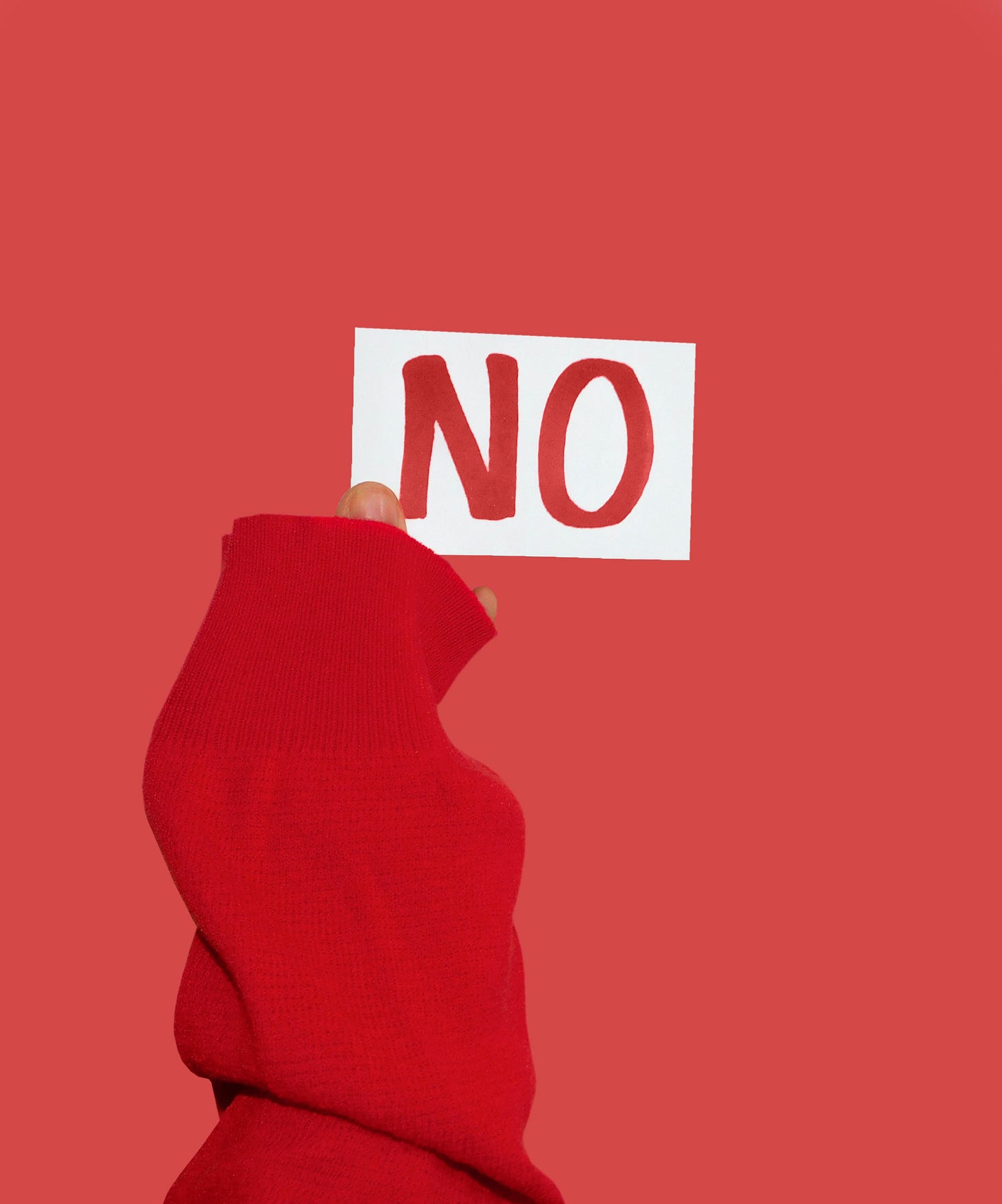 A hand in a read sweater, holds up a white card with the word “no” in red ink, against a red background. A hand in a read sweater, holds up a white card with the word “no” in red ink, against a red background.
