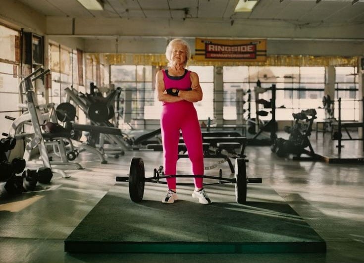 This may contain: an older woman standing on a bench in a gym with weights and equipment behind her