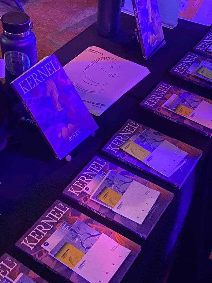Kernel launch party in Gray Area event venue, including contributors in front of a screen, a seated audience at Jasmine's talk, magazines on a table, and various people mingling under purple lighting