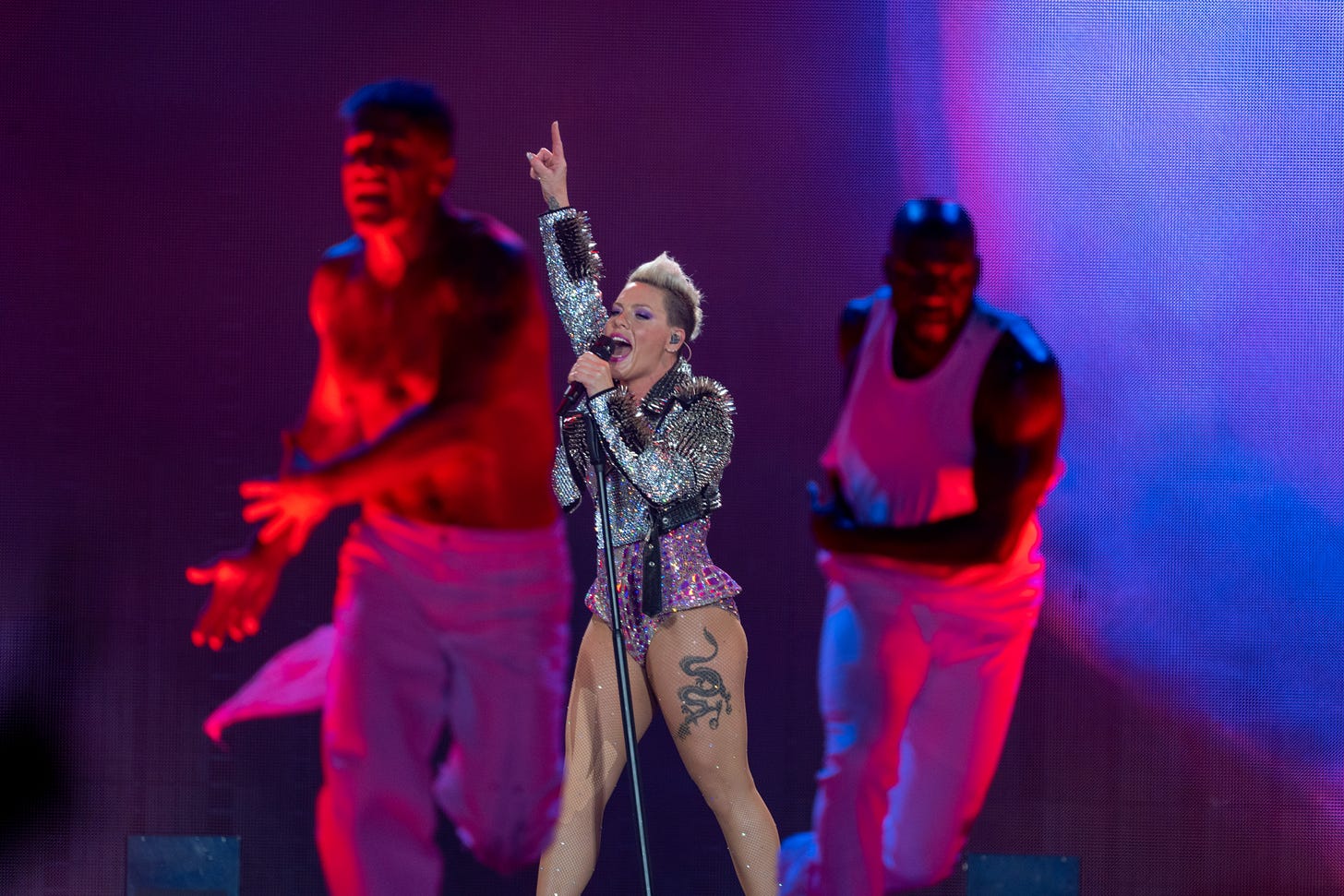 P!nk holds her microphone stand with one hand and points a finger to the sky with the other hand. She is framed between two of her backup dancers, who are illuminated in the foreground in red and blue stage lights.