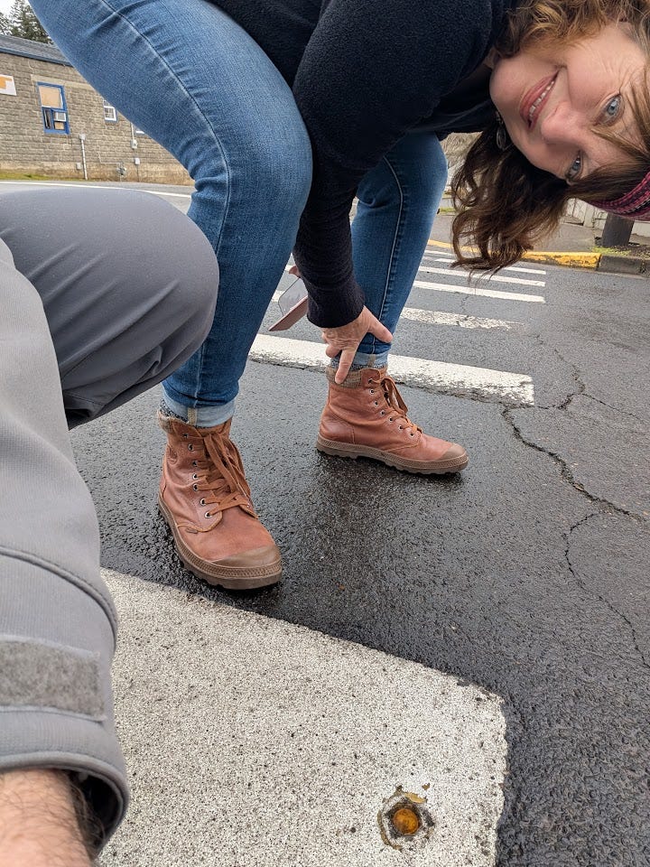 Brown-haired woman in jeans and brown boots upside down and pointing to a penny embedded in the middle of a street