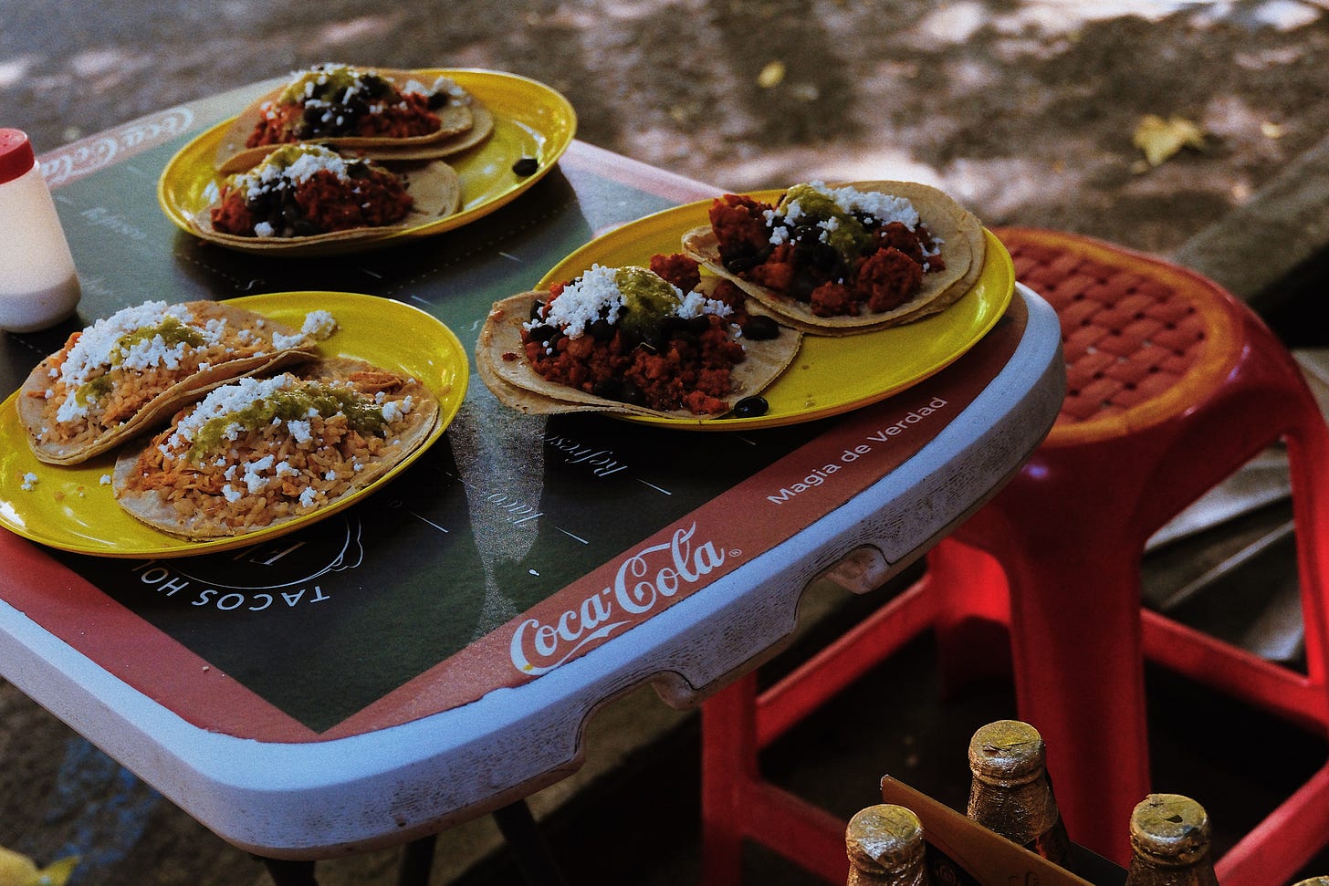 Four plates of tacos topped with salsa verde, queso fresco, and black beans sit on a Coca-Cola branded table next to a red plastic stool at a Mexico City street stall.