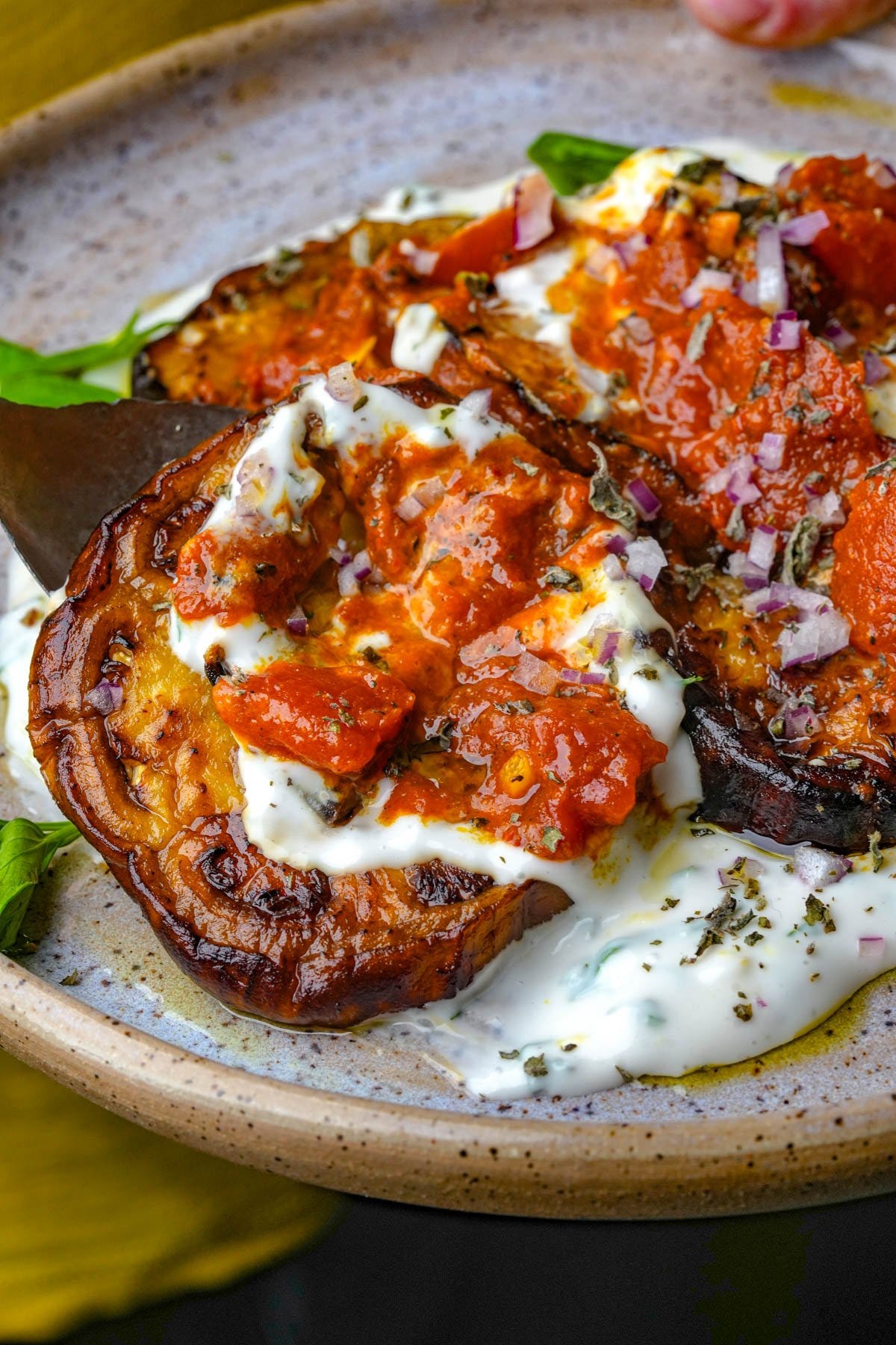 A plate with borani banjan topped with tomato sauce, yogurt, herbs, and finely chopped red onions.