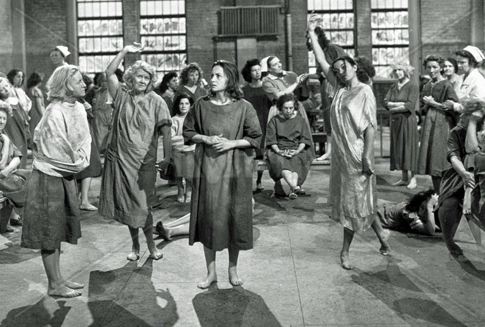Scene from 1948 film The Snake Pit set in a psychiatric hospital. At the centre of a chaotic group of inmates in a large, bare room, Olivia De Havilland stares blankly 