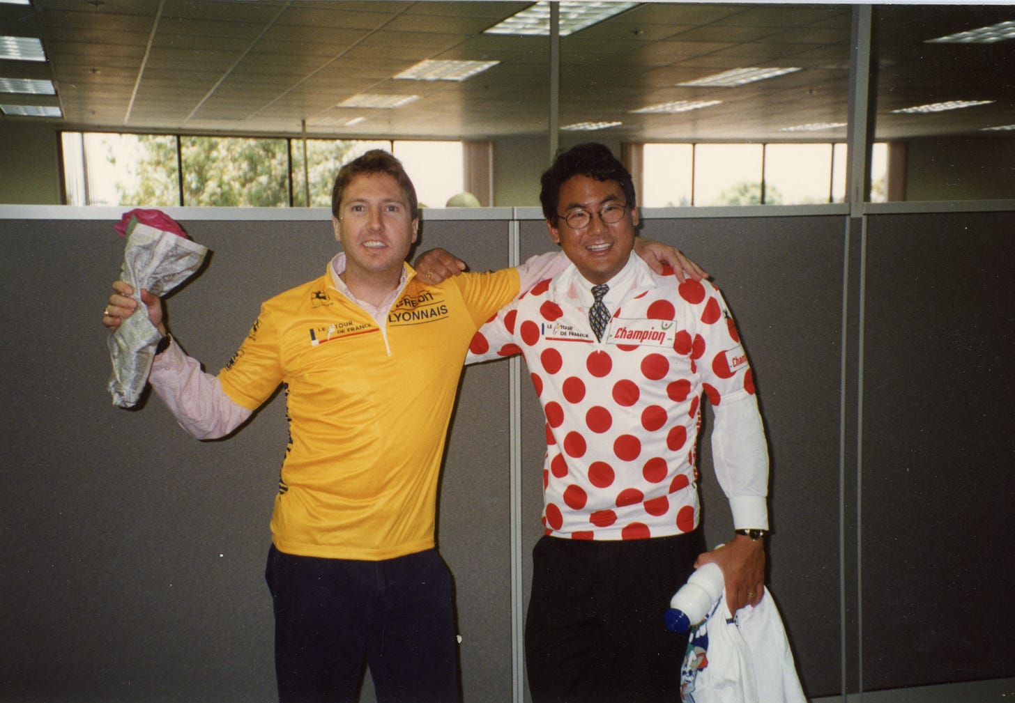 Wayne Fenton and Stephen Pao in a 1990s cubicle office, arms around each other's shoulders and smiling. Fenton wears a yellow Tour de France leader's jersey over a collared shirt, holding a bundled white and pink cloth. Pao wears a red and white polka dot Tour de France jersey over a dress shirt and tie. Wayne Fenton and Stephen Pao in a 1990s cubicle office, arms around each other's shoulders and smiling. Fenton wears a yellow Tour de France leader's jersey over a collared shirt, holding a bundled white and pink cloth. Pao wears a red and white polka dot Tour de France jersey over a dress shirt and tie.