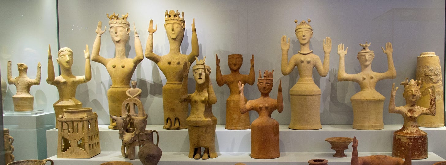 A museum display of terracotta bell jar figurines with upraised arms. The figurines wear different kinds of headdresses, and their arms are in slightly different positions - some with the palms facing forward, some with the palms facing inward, some with the arms directly out to the side and some with the arms slightly forward. Three of the figurines also have adorable little feet sticking out from the bottom of the skirt.