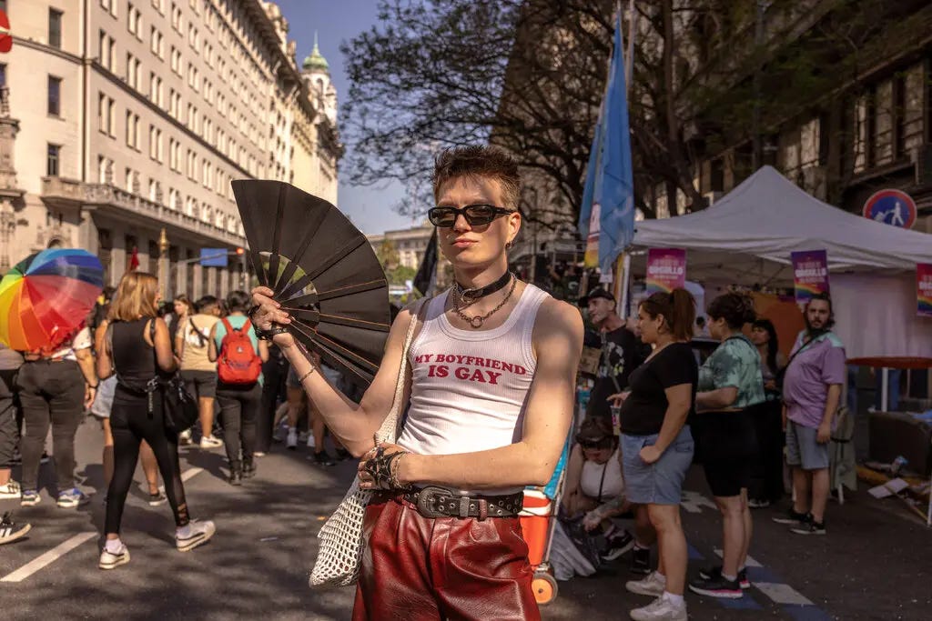 A man in narrow sunglasses waving a black fan and wearing at tank top that reads “My Boyfriend is Gay.”