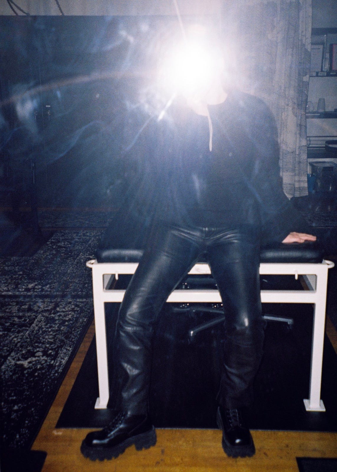 A film mirror selfie of Evyn Silver, a white femme wearing a black sweater, black leather pants, and chunky black leather boots. She's leaning against a metal table with a leather surface and in the background are metal cabinets. The flash from the camera obscures her face.