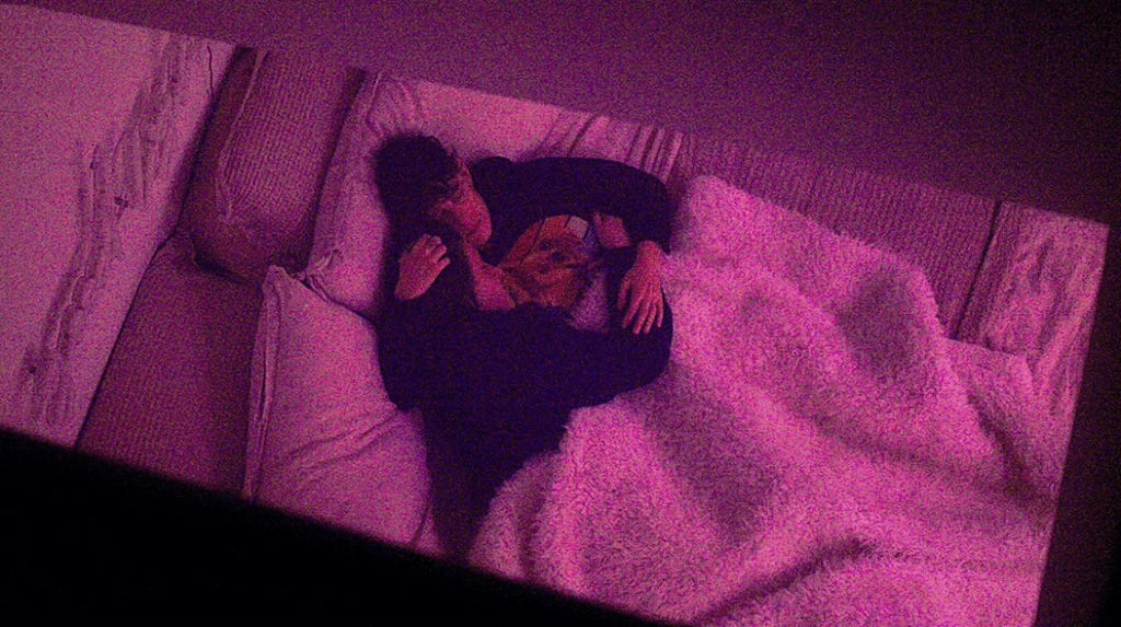 An aerial shot of Andrea holding Meg in their bed, as taken from their roof, framed by a sliver of their sky-light.