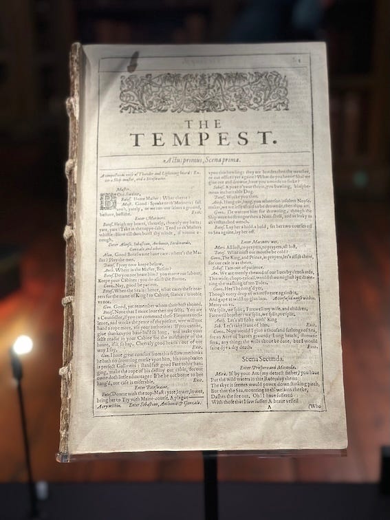 Photograph of a page from an early printed book taken through the glass of a display case. Cover has been ripped off; page has the title 'The Tempest' in large letters with decorative foliage printed above then two columns of densely printed text below. 