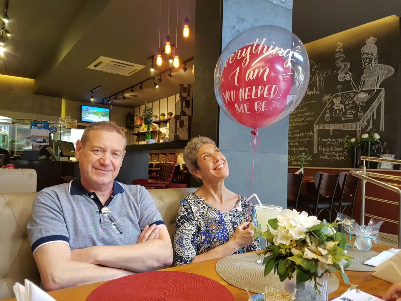 A white woman with white hair and a white man with blond hair, both in their 60s, sit in a restaurant booth. The woman holds a ballon. A white woman with white hair and a white man with blond hair, both in their 60s, sit in a restaurant booth. The woman holds a ballon.