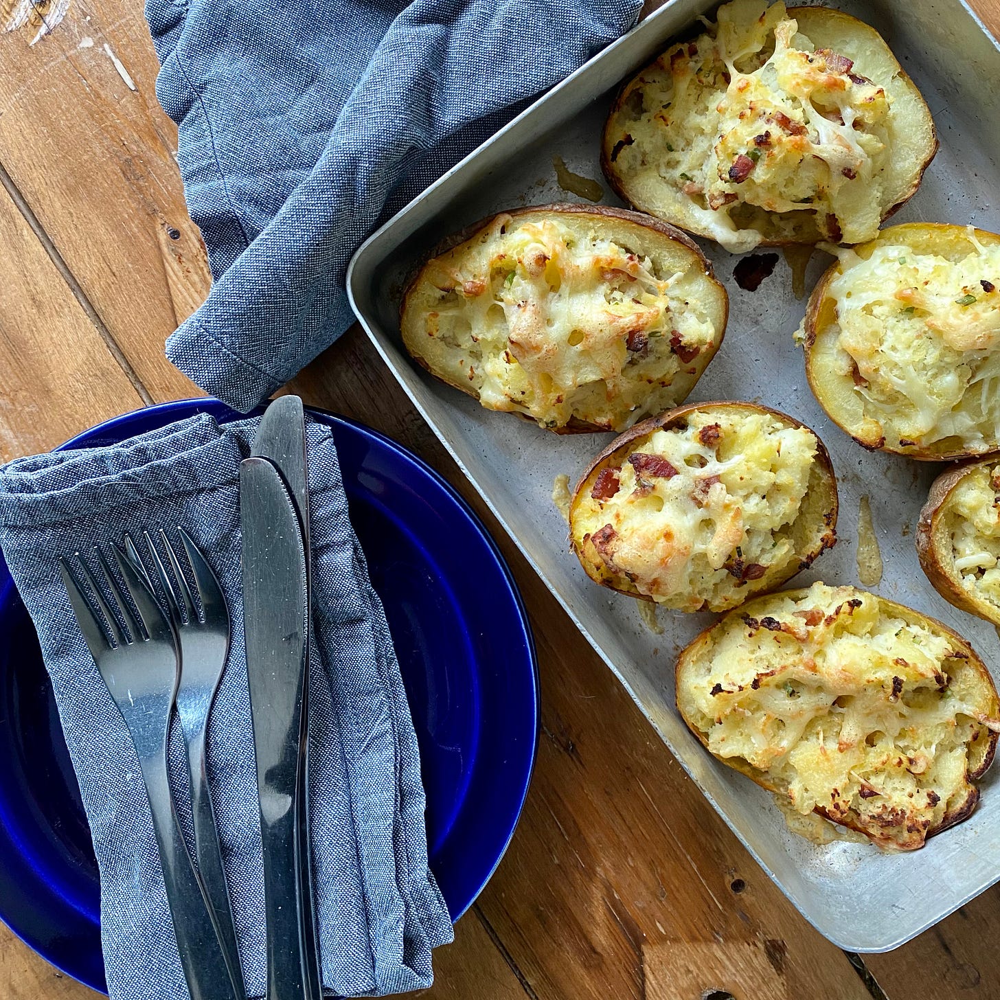 Oven tray with filled backed potatoes, next to a blue plate with a napkin and cutlery