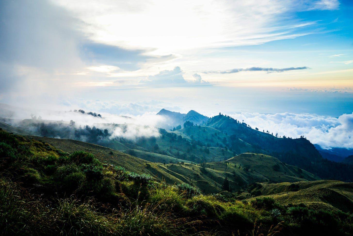 A green mountain landscape covered with mist and clouds under a bright blue sky in Indonesia.