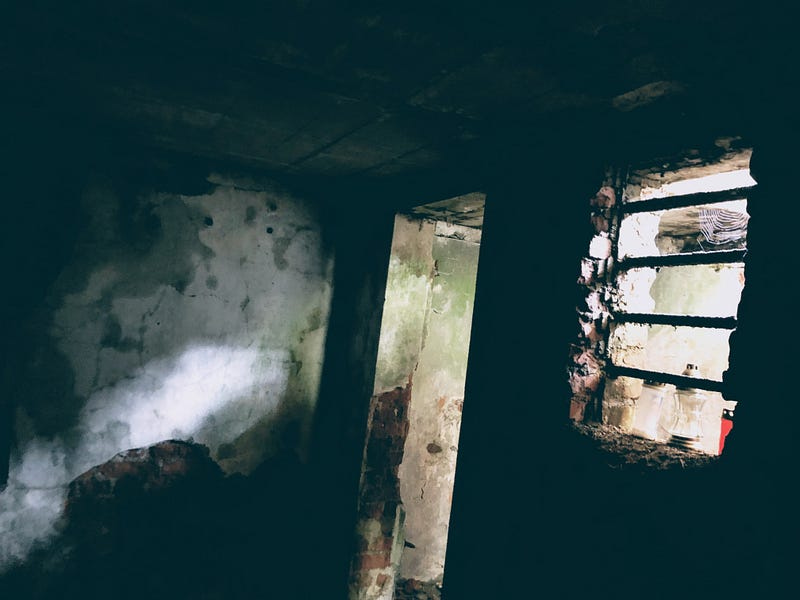 Colored picture of the inside of the underground correctional cell at the location of the Jáchymov forced labor camp. The walls are decrepit and faint lights penetrates from the door frame and the window with horizontal bars on the right side. Some candle holders are visible on the outside of the window.
