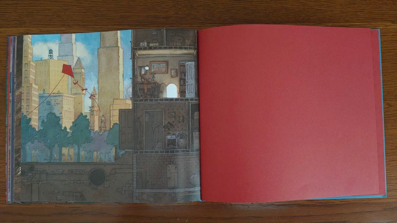 Doppia pagina del libro "Viaggio" di Aaron Becker appoggiata su un tavolo di legno marrone. A sinistra l'illustrazione di uno scorcio cittadino di grattacieli, un aquilone rosso che vola e la sezione di una casa. A destra pagina rossa.