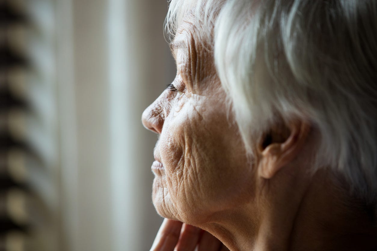 Profile of white older woman with gray hair and wrinkled face against light-colored background.