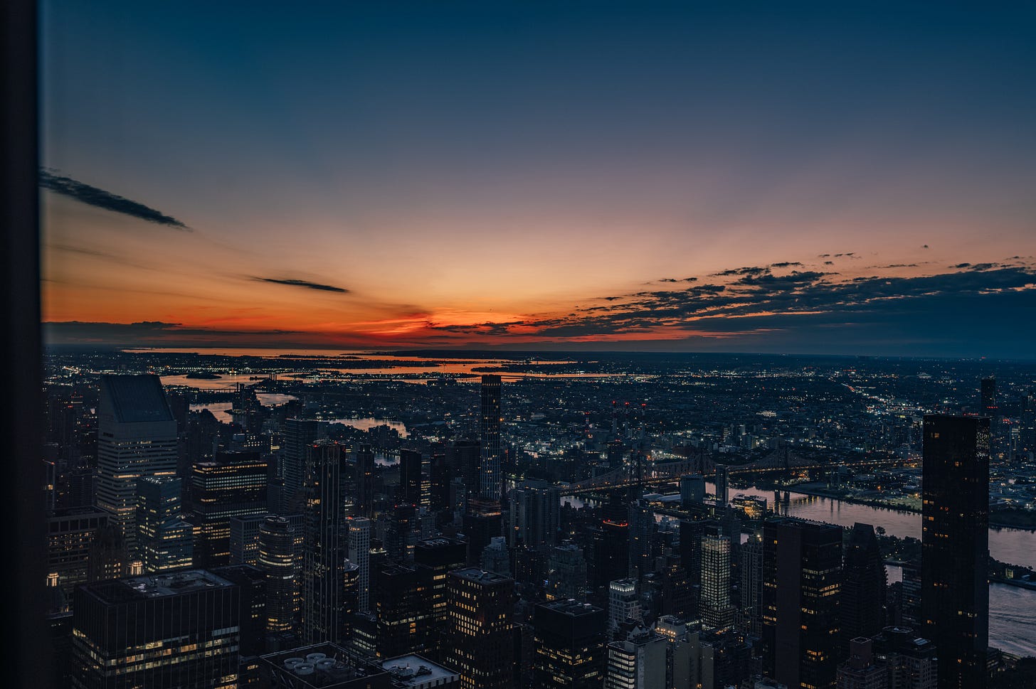 A wide pre-sunrise view over Manhattan from high above, the city still lit below, a long orange band along the horizon and dark cloud streaks above it. A wide pre-sunrise view over Manhattan from high above, the city still lit below, a long orange band along the horizon and dark cloud streaks above it.