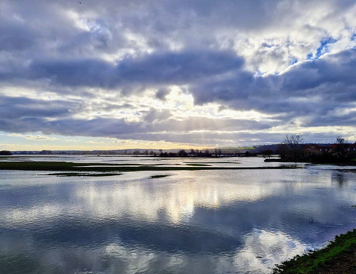 A pub garden terrace flooded with water with the river and a small bridge in the background; a vast expanse of water dotted with islands reflecting a cloudy sky