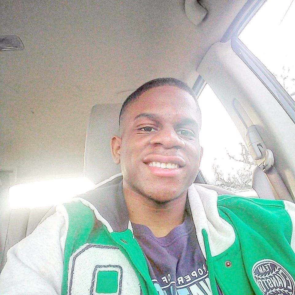 A man sitting in a car smiles and looks into the camera while wearing a green jacket with white sleeves.