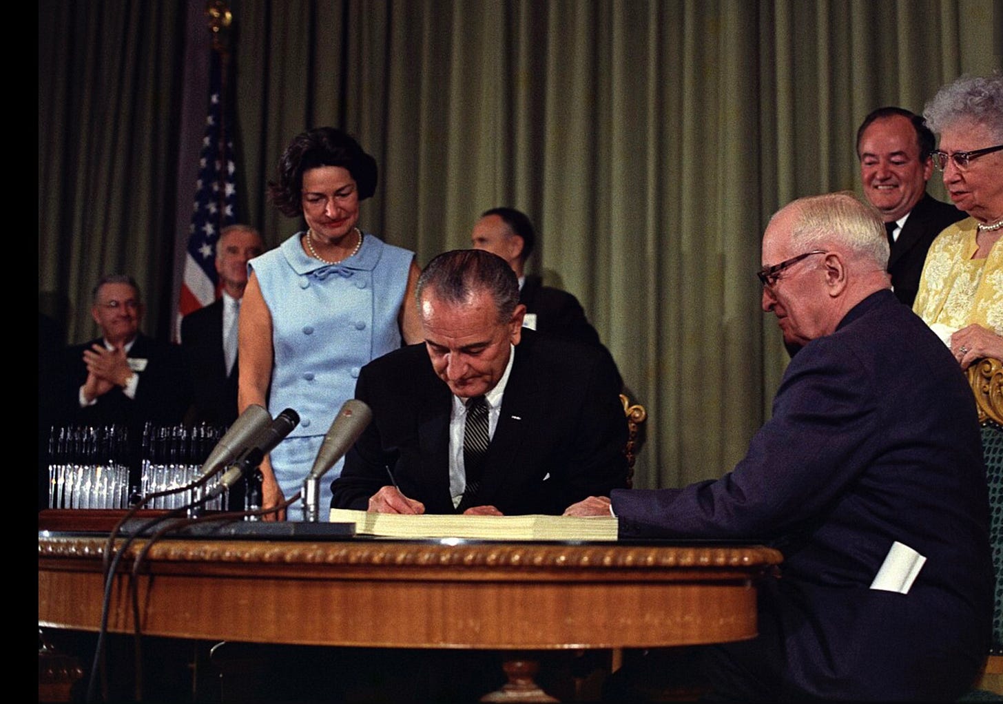 Women in summer dresses and men in suits and ties stand reverently around Johnson as he signs a bill into law.