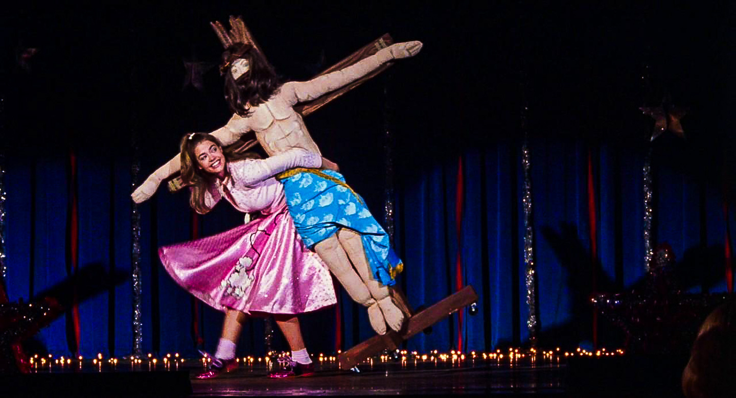 A smiling teenage girl in a pink poodle skirt joyfully dances with a life-size crucifix of Jesus on a blue-lit stage, surrounded by twinkling lights and tinsel decorations. A smiling teenage girl in a pink poodle skirt joyfully dances with a life-size crucifix of Jesus on a blue-lit stage, surrounded by twinkling lights and tinsel decorations.